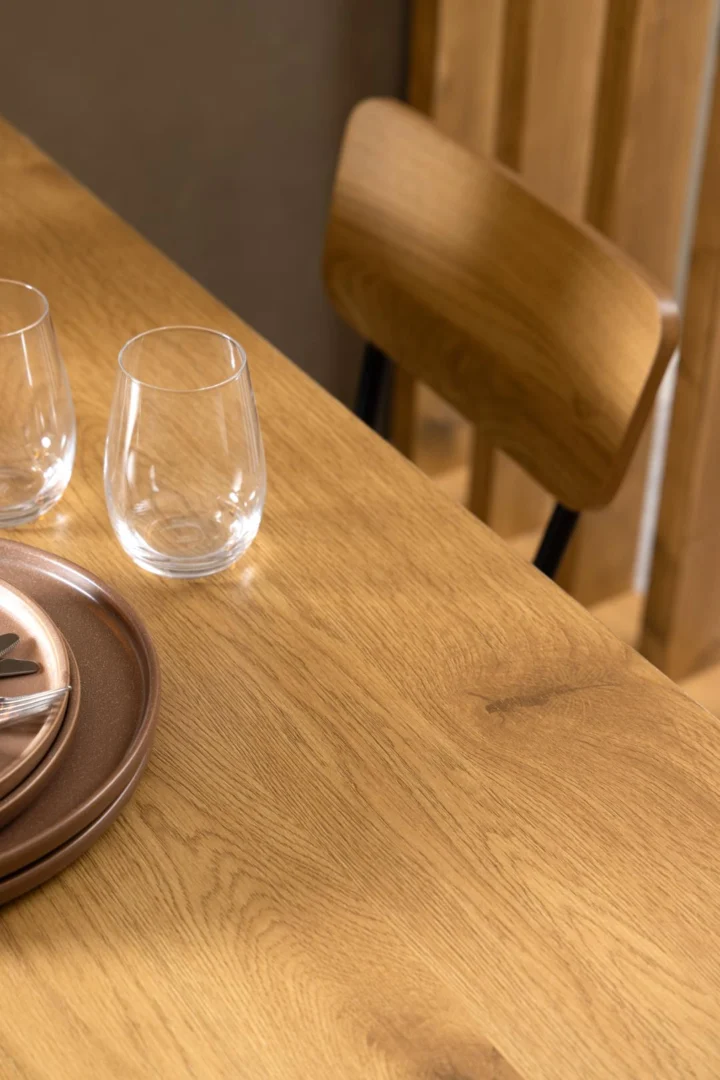 Een warme, minimalistische setting met de Eettafel Nieuwe Sfeer 80 x 75 CM Rond in eikenhout staal, set met twee heldere stemloze glazen, gestapelde bruine borden en een vork naast een bijpassende houten stoel met gebogen rug.