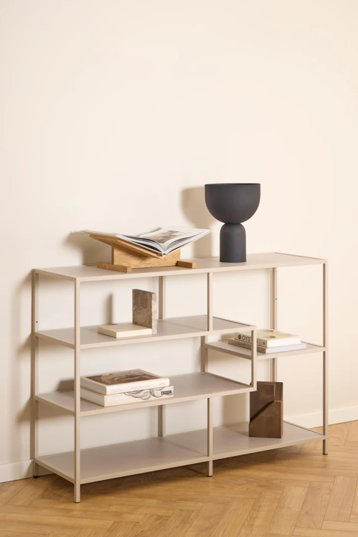 De Seaford boekenkast, een minimalistische beige metalen boekenplank, toont boeken in verschillende schikkingen, een open boek op een houten standaard en een strakke zwarte vaas tegen een lichte muur en een vloer van licht hout met visgraatmotief.