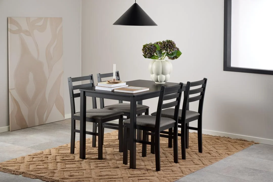 In een moderne eetkamer staat de Eettafel Nieuwe Sfeer 110 x 70 CM (rechthoekig MDF), een set voor vier personen met grijze stoelen met kussens, een beige vloerkleed met structuur, een vaas met bloemen, boeken en een kaars onder een zwarte hanglamp in neutrale tinten.