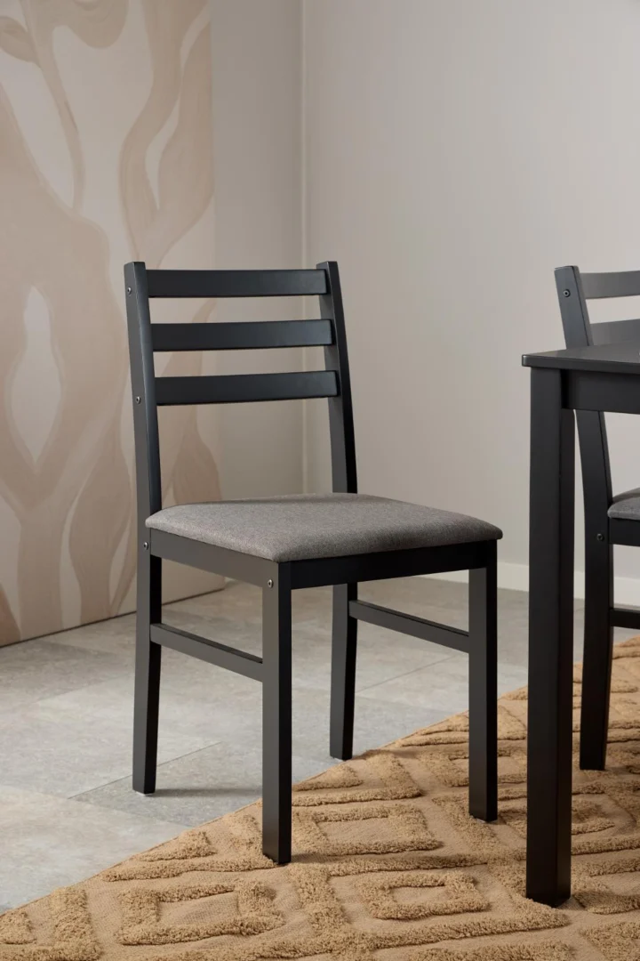 Een zwarte houten stoel met een grijs kussen staat naast de Eettafel Nieuwe Sfeer 110 x 70 CM (rechthoekig MDF) op een beige vloerkleed met structuur in een moderne, minimale kamer met lichte muren en betegelde vloeren.