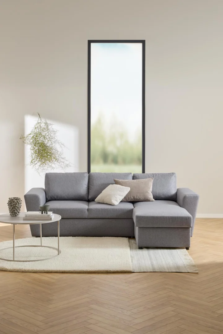 Een moderne woonkamer met een grijze sectionele bank, lichte kussens, een Salontafel Grijs 80 CM Rond Kunststof met decor, een wit vloerkleed op een houten vloer en een groot raam met vaag groen buiten.