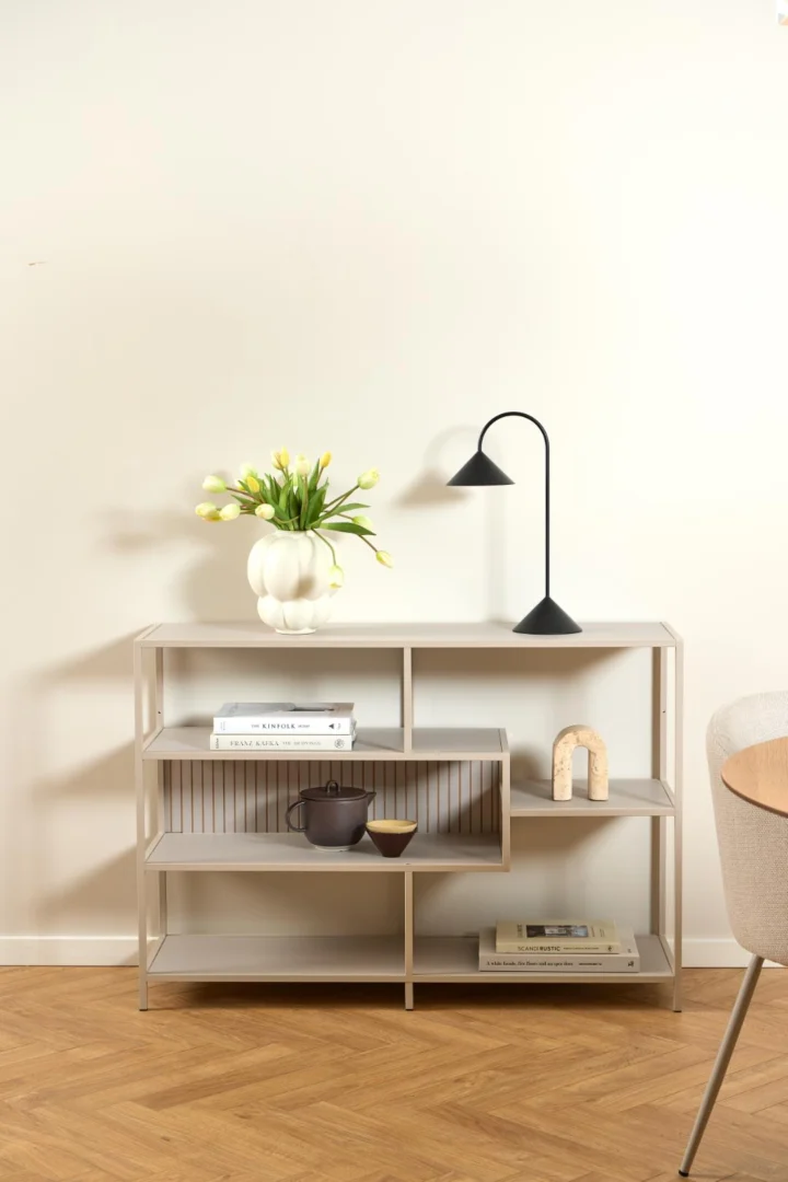 De Seaford boekenkast, een moderne witte plank, toont gele tulpen in een vaas, een zwarte lamp, boeken, een kleine boog, een theepot en een schaal. Het staat tegen een effen witte muur met een houten vloer en een beige stoel die gedeeltelijk zichtbaar is.