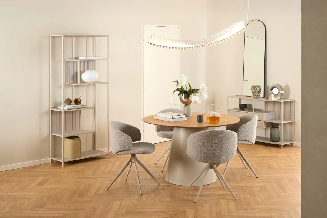Een moderne eetkamer met een ronde houten tafel, vier grijs gestoffeerde stoelen, een gebogen armatuur, een hoge spiegel, de Seaford boekenkast voor open kasten en decoratieve items op een lichte houten vloer.