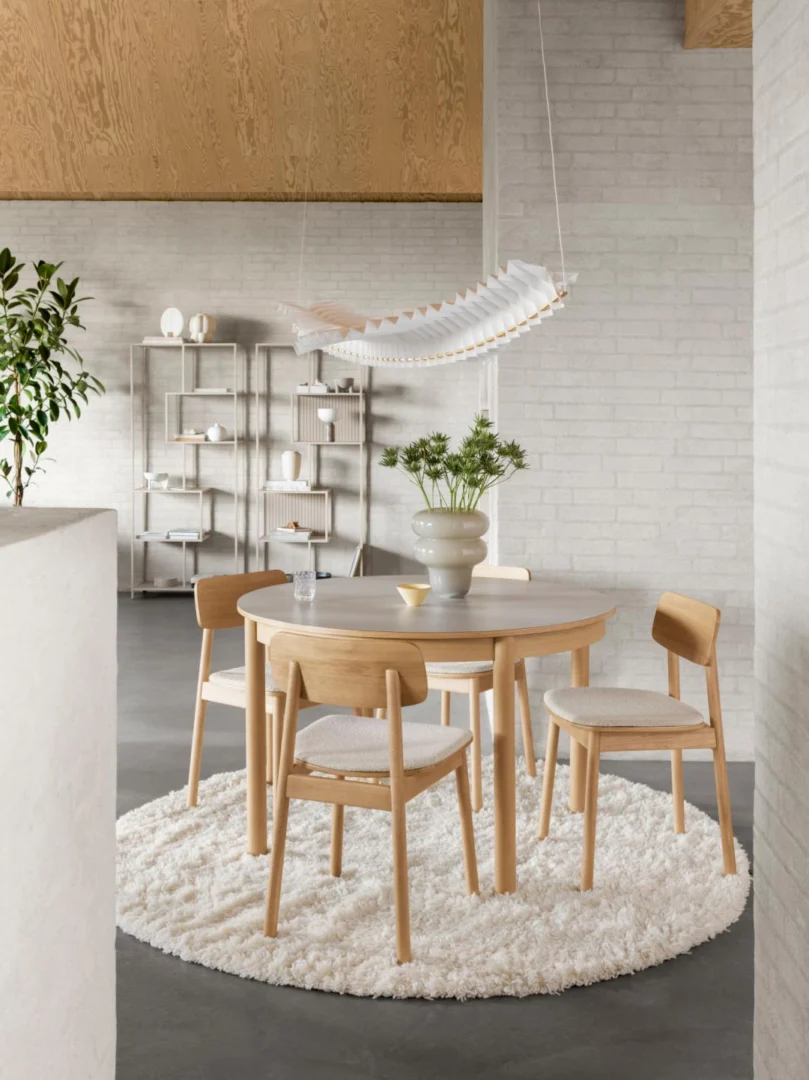 Een moderne eethoek bestaat uit een ronde houten tafel met vier bijpassende stoelen op een wit gestructureerd tapijt. Boven de tafel hangt een sculpturale hanglamp en op de achtergrond staat de Seaford boekenkast met decoraties en een potplant.