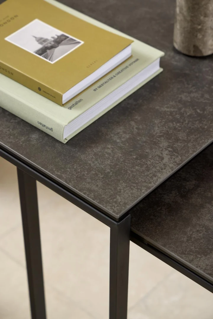 Een close-up van de Salontafel Zwart 50 x 55 CM Vierkant, een kunststof tafel met donkere structuur en zwarte metalen poten, met groene en gele hardcover boeken erop gestapeld en een deel van een cilindervormig voorwerp zichtbaar in de hoek.