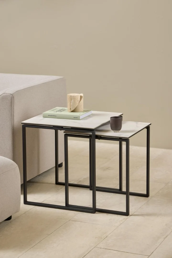 Naast een lichtgrijze bank staan twee Salontafel Zwart, Wit 50 x 55 CM Vierkant tafels gemaakt van kunststof, met zwarte frames en witte bladen. Op de tafels staan een beige mok, een groen boek en een klein donker kopje. De vloer is van lichte tegels.