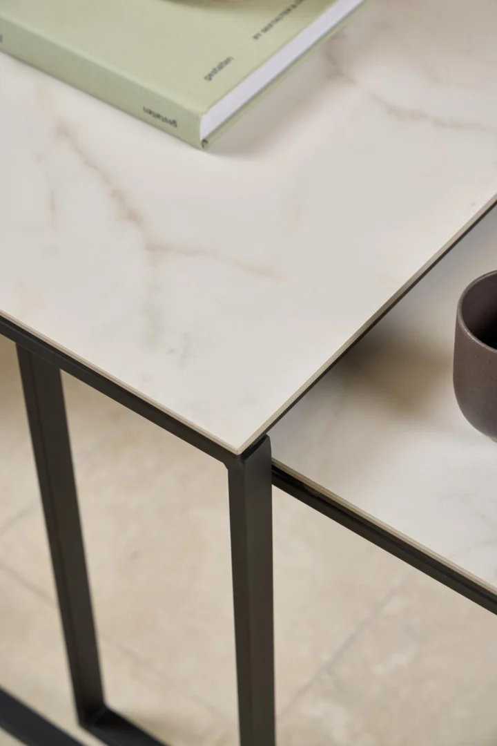 Close-up van de Salontafel Zwart, Wit 50 x 55 CM Vierkant met een dun, lichtgekleurd kunststof blad, zwarte poten, een groen boek en een klein bruin kopje op het oppervlak.