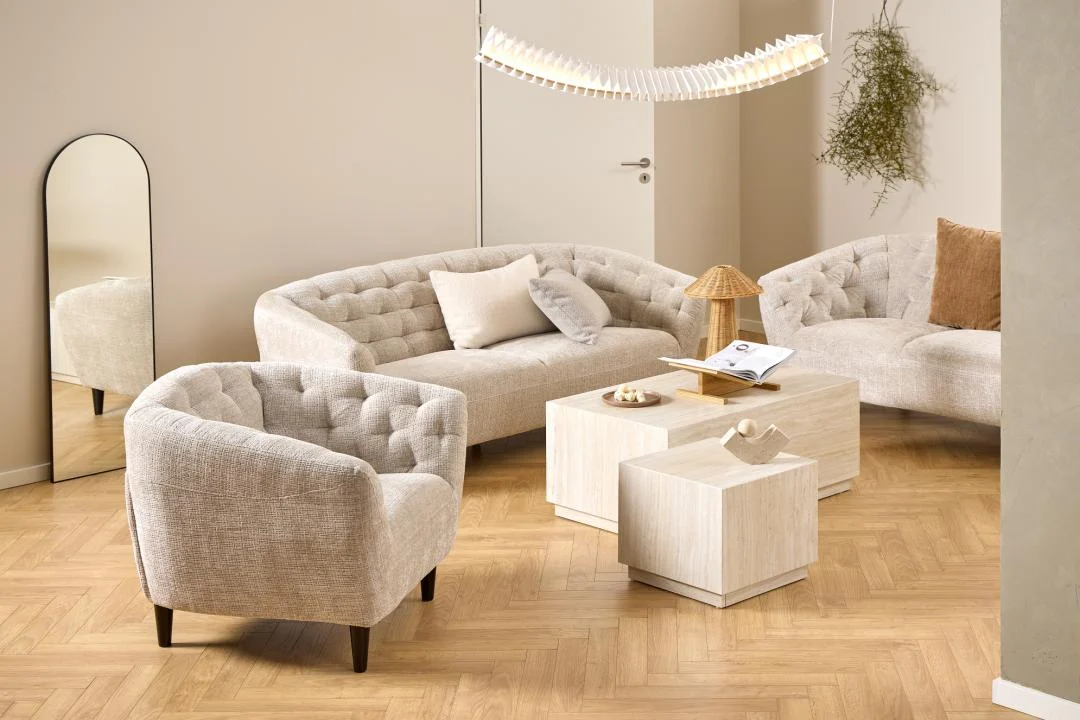 Een moderne woonkamer met beige getufte banken, een bijpassende fauteuil, twee Salontafel Beige Roest Rechthoek kunststof salontafels, een geweven lamp, een gebogen vloerspiegel en een hangplant op een vloer van visgraathout.