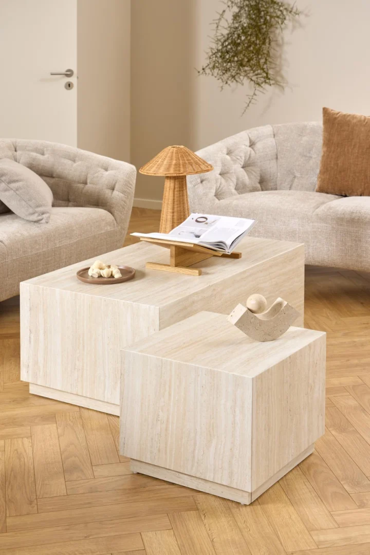 In een moderne woonkamer staan twee Salontafel Beige, Roest Rechthoek salontafels van kunststof, met een boek, kleine lamp en decor erop. Op de achtergrond staan gebogen beige banken en een bruin kussen.