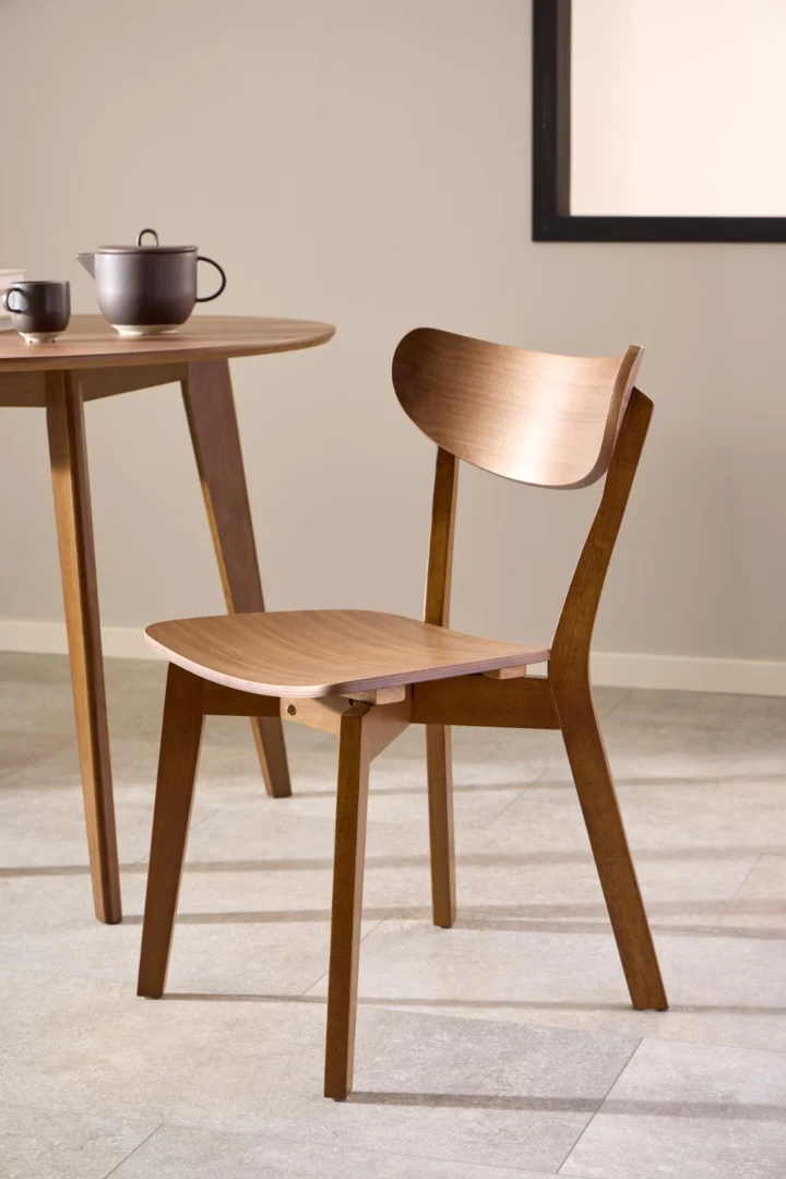 De Eettafel Nieuwe Sfeer 105 CM Rond Mdf Massief, set met een theekopje en theepot, staat naast een houten stoel met gebogen rugleuning op een lichte tegelvloer in een minimalistische kamer.