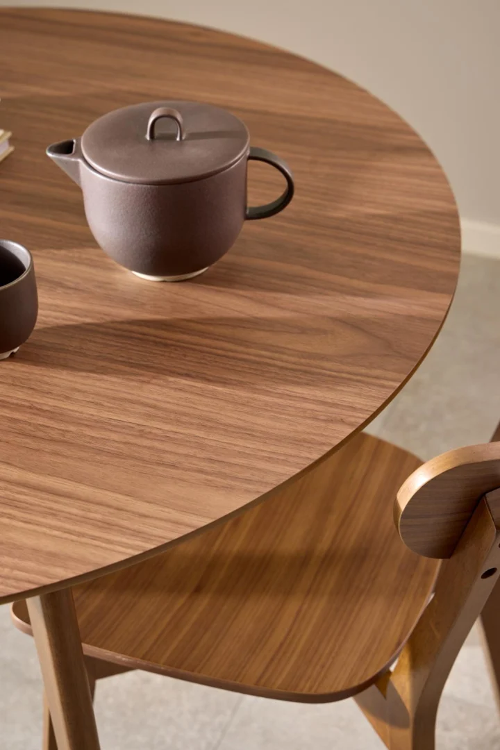 De Eettafel Nieuwe Sfeer 105 CM Rond MDF Massief is gecombineerd met een bijpassende houten stoel. Op de tafel staan een bruine theepot en beker van keramiek, waardoor een minimale, moderne setting in neutrale tinten ontstaat.