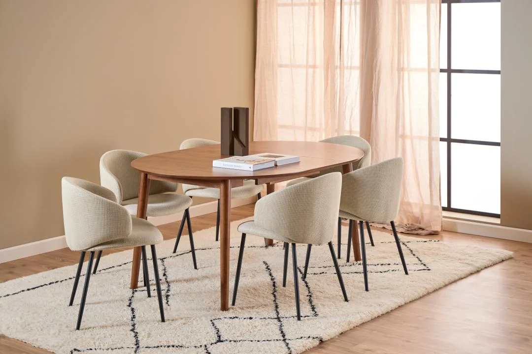 Een moderne eetkamer met de Eettafel Nieuwe Sfeer 115 CM Rond uitschuifbaar in notenhout, zes beige stoelen en een vloerkleed met geometrisch patroon. Het licht valt door de transparante gordijnen en creëert een warme, uitnodigende sfeer.