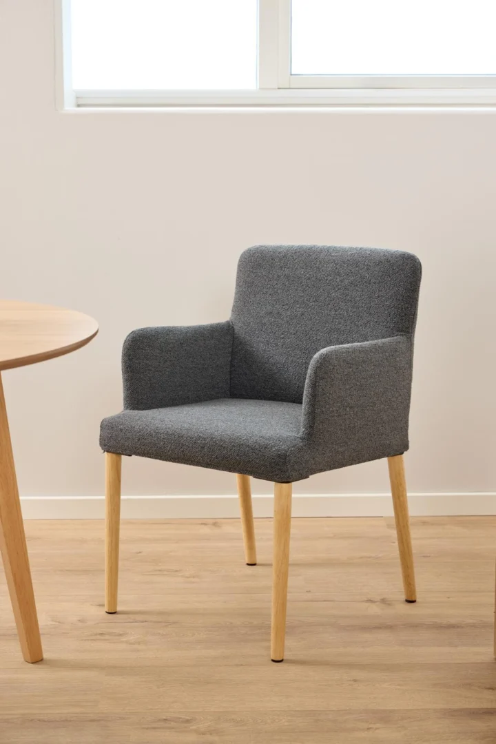 Een modern grijs gestoffeerde fauteuil met lichthouten poten staat op een lichthouten vloer naast de Eettafel Nieuwe Sfeer 120 CM Rond, een ronde tafel van eikenhout, met een witte muur en raam op de achtergrond.