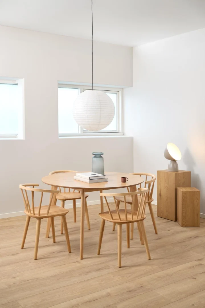 Een minimalistische eethoek met de Eettafel Nieuwe Sfeer 120 CM Rond in eikenhout, vier bijpassende stoelen, een papieren lantaarn hanglamp en gestapelde houten kubussen met een vaas en moderne lamp. Lichthouten vloeren en witte muren maken de look compleet.