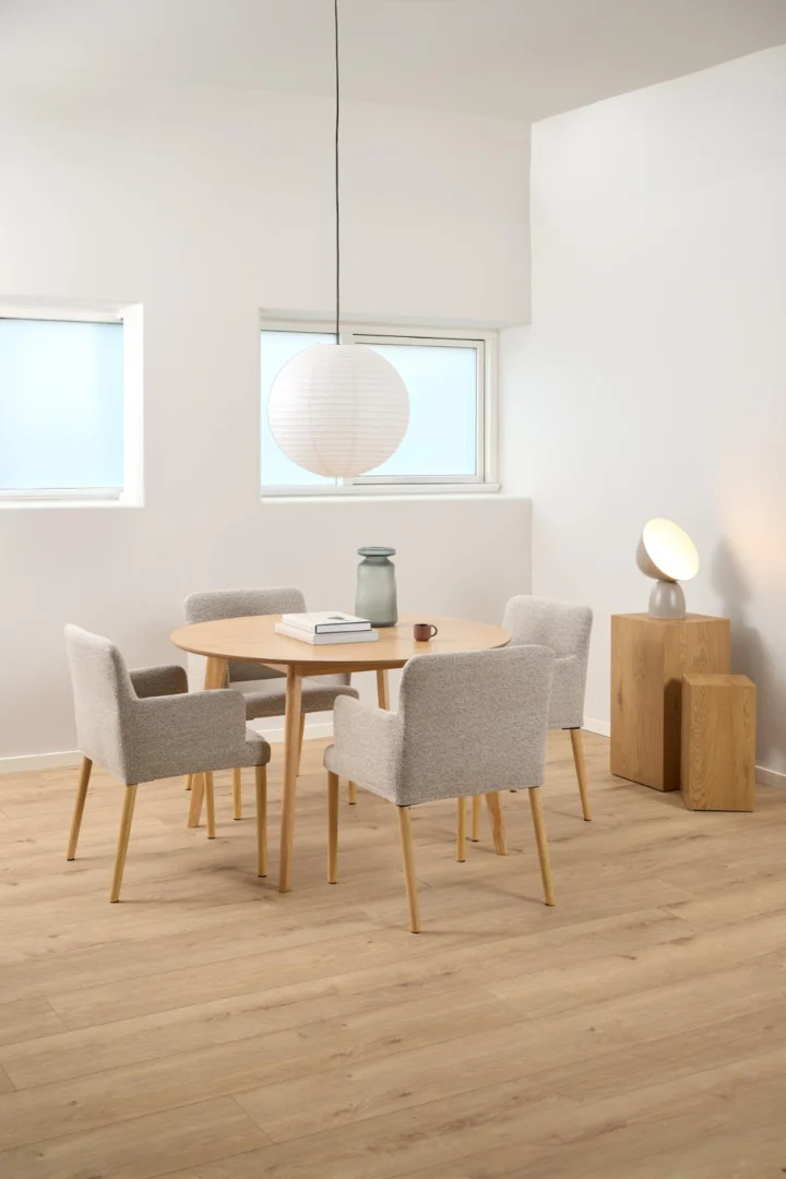 Een moderne eethoek met de Eettafel Nieuwe Sfeer 120 CM Rond in eikenhout, omringd door vier beige gestoffeerde stoelen, met een witte papieren hanglamp, lichte houten vloeren en minimale decoratie zoals een vaas en een sculpturale lamp.