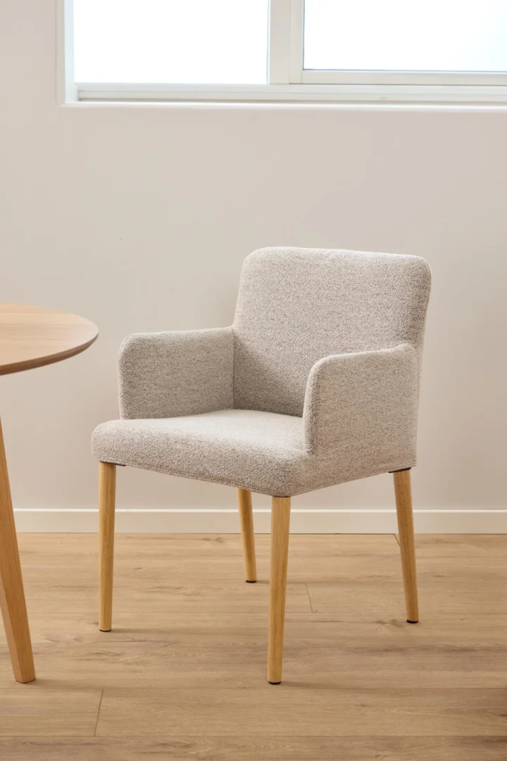 Een modern gestoffeerde fauteuil met lichtgrijze stof en houten poten staat bij de Eettafel Nieuwe Sfeer 120 CM Rond, gemaakt van eikenhout, op een lichthouten vloer met een raam erboven.