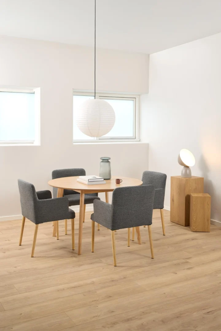 De Eettafel Nieuwe Sfeer 120 CM Rond, gemaakt van eikenhout, is gecombineerd met vier grijs gestoffeerde stoelen in een minimalistische kamer met een licht houten vloer, een hanglamp met papieren lantaarn en een bijzettafel met lamp en decor.