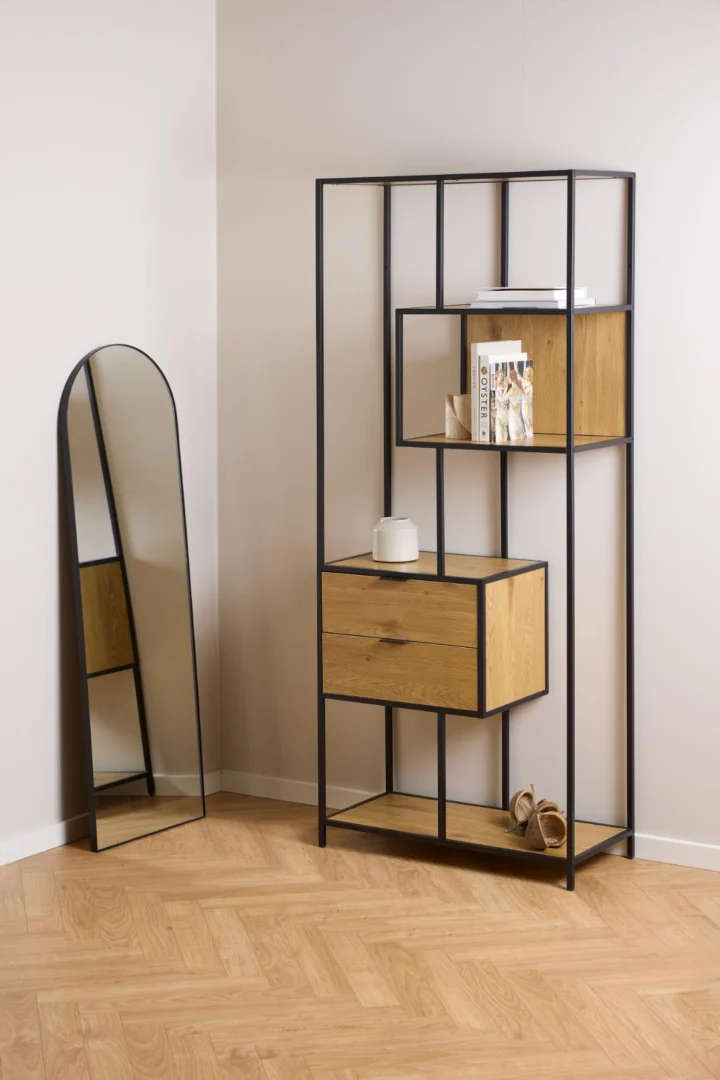 Zijaanzicht van een zwarte metalen boekenkast met wild eiken accenten en twee lades, op een houten vloer. Een spiegel staat ernaast.