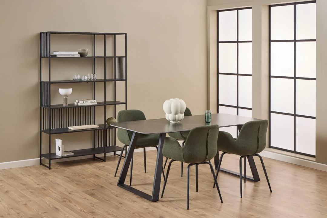 Een moderne eetkamer met een rechthoekige tafel, zes groene stoelen, een decoratief middenstuk, houten vloeren, grote rasterramen met natuurlijk licht en een zwart metalen Strington boekenkast met verschillende items.