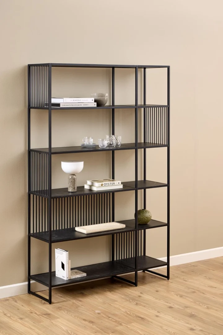 De Strington boekenkast, een zwart metalen boekenplank met vijf planken, toont boeken, glasdecor, een witte lamp, een kleine plant en een beige schaal tegen een lichtbeige muur op een houten vloer.