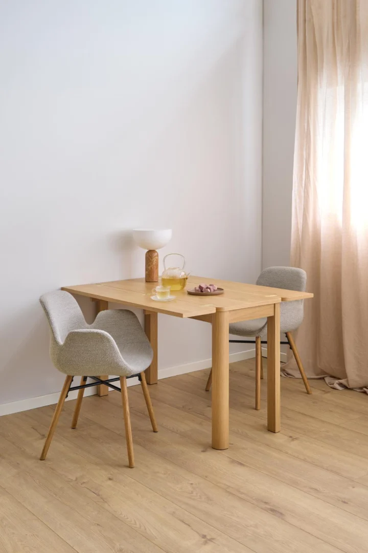 De Eettafel Nieuwe Sfeer (90 x 76 CM, rond, eikenhout) is gecombineerd met twee lichtgrijs gestoffeerde stoelen in een minimalistische kamer. Op tafel staan een glazen theepot, kopjes, houten vaas en witte lamp. Lichtbeige gordijnen bedekken een deel van het raam.
