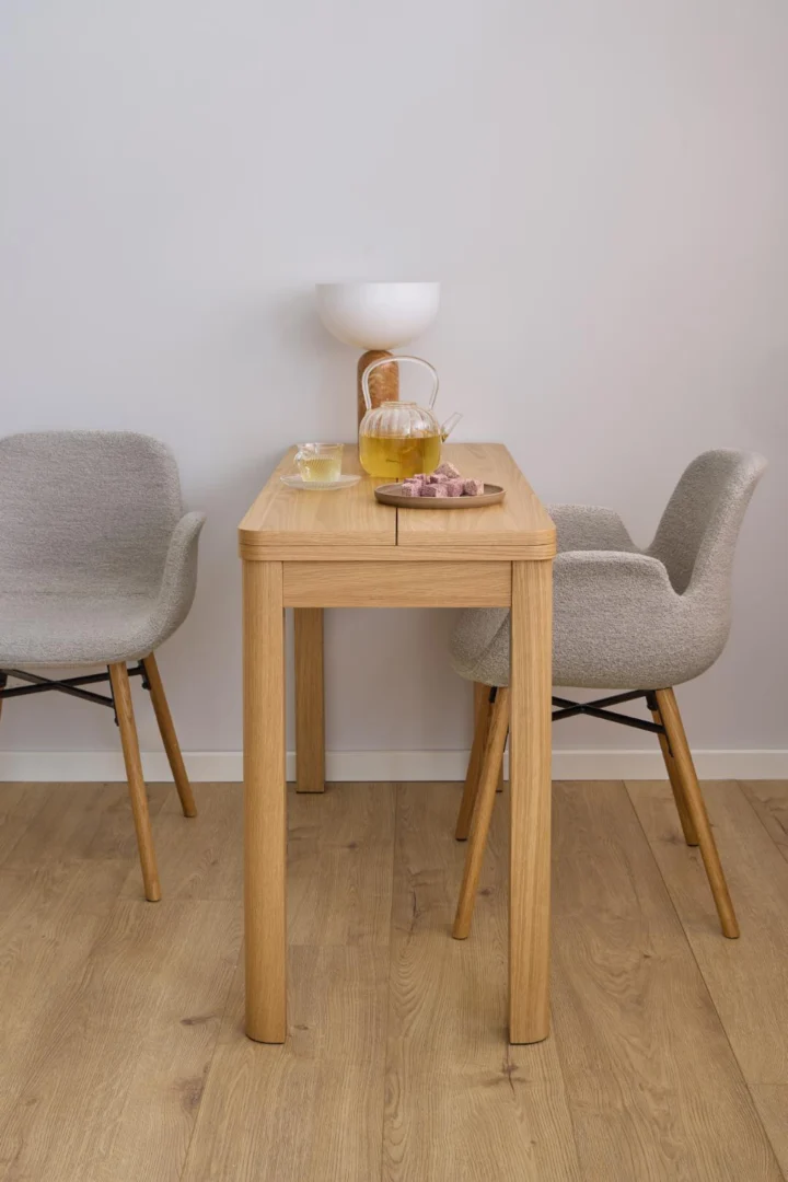 Een minimalistische eettafelopstelling met de Eettafel Nieuwe Sfeer 90 x 76 CM Rond in eikenhout, twee grijze stoelen met kussens, een glazen theepot met kruidenthee, twee glazen, een snackschaal en een witte kom, tegen een effen witte muur.