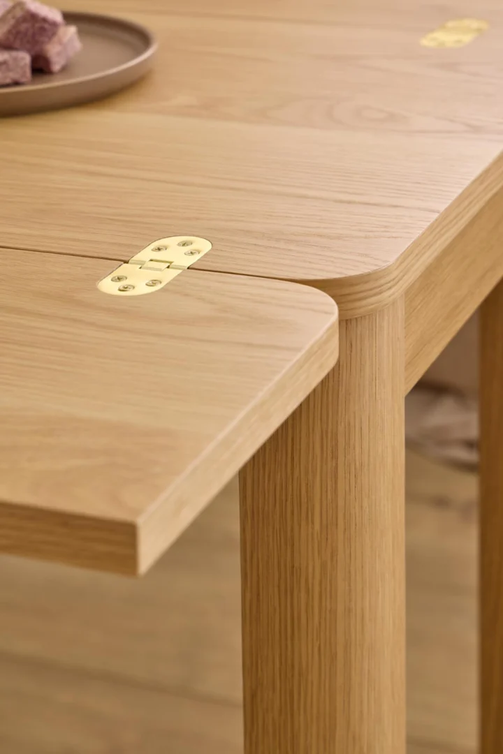 Een close-up van de Eettafel Nieuwe Sfeer 90 x 76 CM Rond, gemaakt van eikenhout met afgeronde hoeken en zichtbare messing scharnieren. Een klein bordje met etensresten verschijnt gedeeltelijk in de linkerbovenhoek.