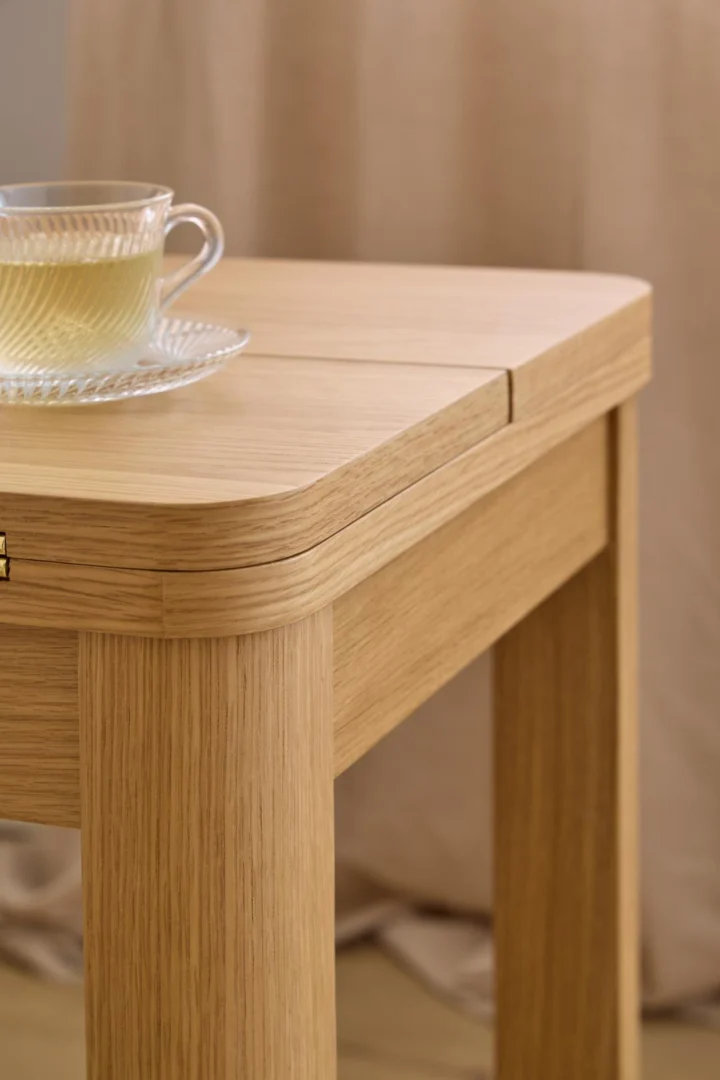 Een glazen kop en schotel met lichtgekleurde thee zit op de hoek van een Eikenhout Eettafel Nieuwe Sfeer 90 x 76 CM Rond. Op de achtergrond hangt een beige gordijn.