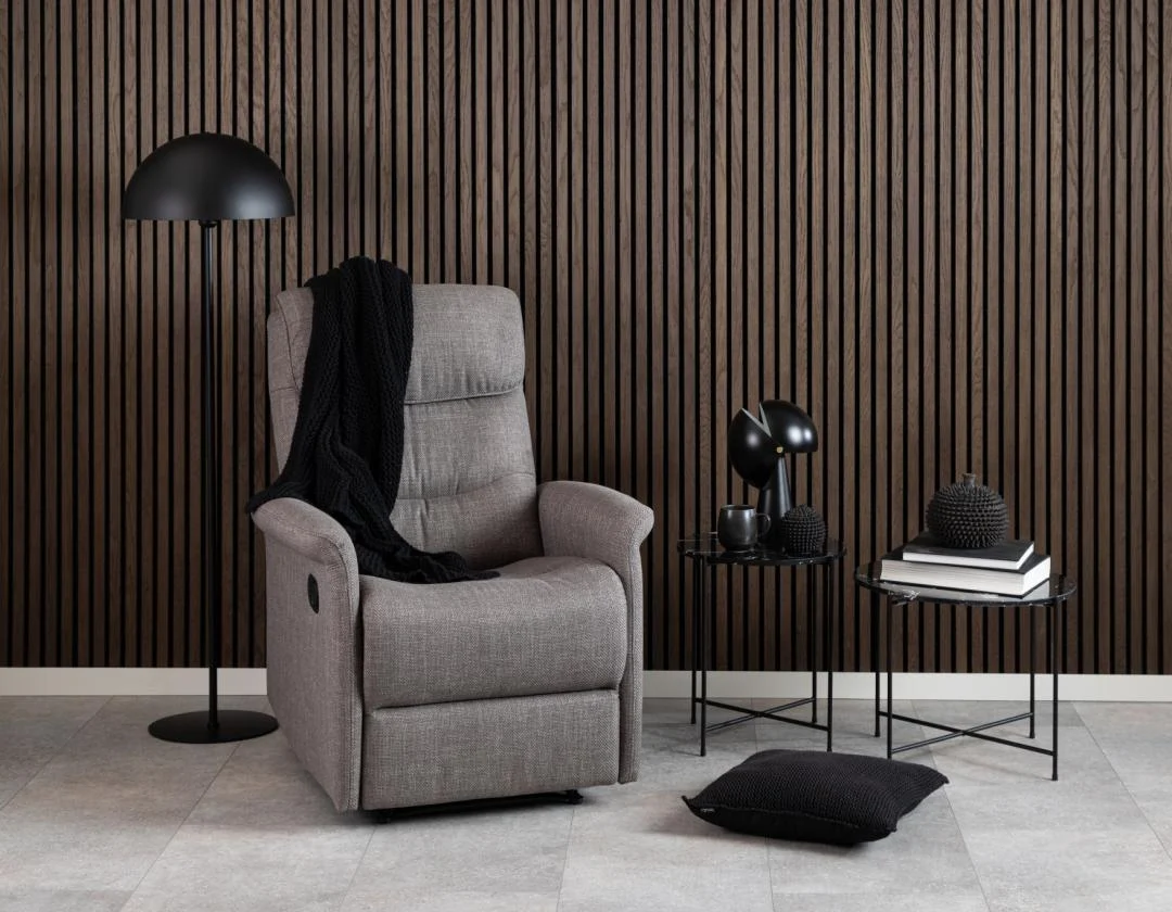 Een moderne leefruimte met een grijze relaxfauteuil, zwarte staande lamp, Bijzettafel Zwart 42 CM Rond Kunststof met gestapelde boeken, zwart decor, een zwart gebreid kleed en kussen, alles tegen een donkerhouten lattenwand.