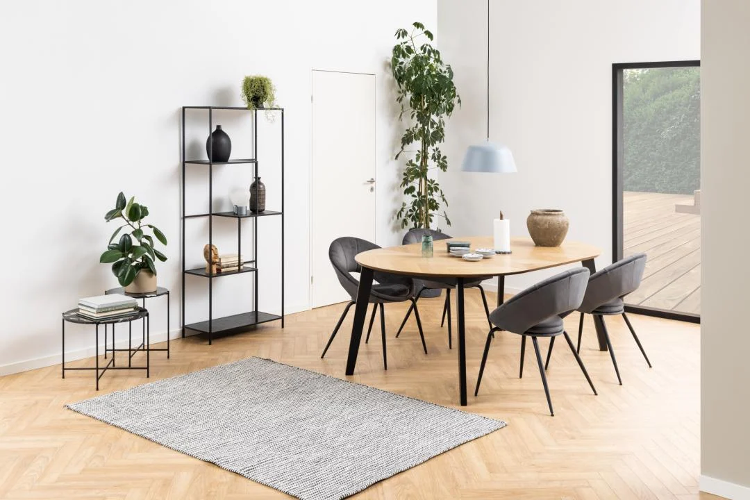 Moderne eetkamer met een houten ovale tafel, vier zwarte stoelen, de Bijzettafel Zwart 42 CM Rond Kunststof, zwarte planken, kamerplanten, een hanglamp, een groot raam naar een houten terras, lichte houten vloeren en een grijs vloerkleed.