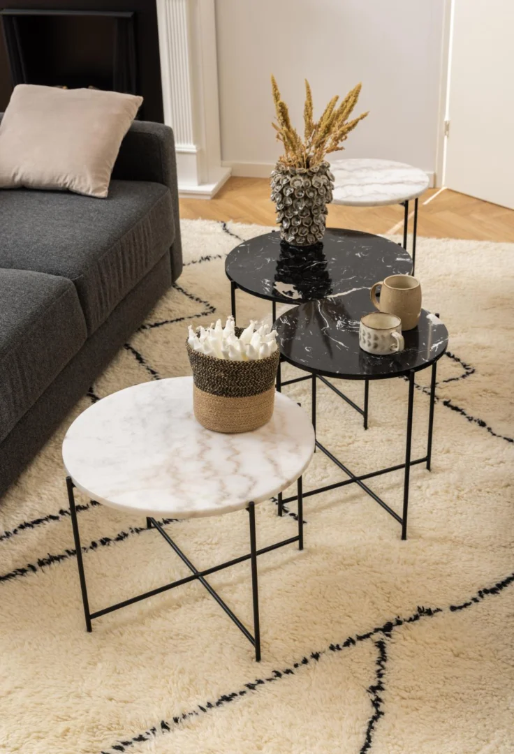 Drie Bijzettafel Zwart 42 CM Rond Kunststof tafels met zwarte metalen poten staan op een vloerkleed met crèmekleurig patroon bij een grijze bank, met decor zoals een vaas met gedroogde tarwe, mokken en een mand met witte voorwerpen.