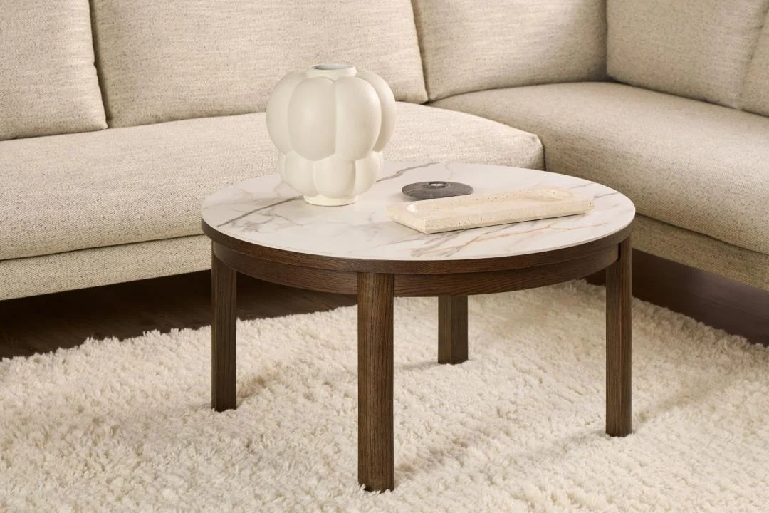 Een Salontafel Beige 90 CM Rond Eikenfineer ligt op een wit vloerkleed met structuur, met daarop een sculpturale witte vaas, een rechthoekig voorwerp en een klein rond voorwerp. Een beige sofa verschijnt op de achtergrond.
