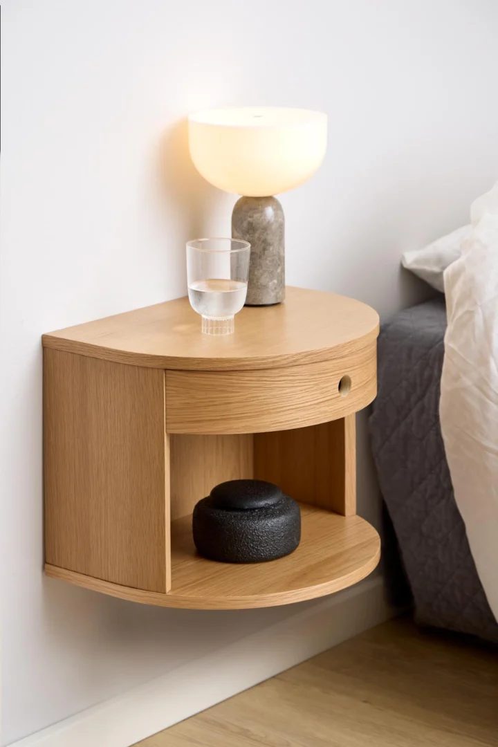 Het Nachtkastje Eikenhout 40 x 30 x 31 CM, een houten zwevend nachtkastje met een halfrond ontwerp, lade en plank, is gemonteerd op een witte muur naast een bed met een wit dekbed. Bovenop staan een glas water en een lamp; op de plank staat een zwart voorwerp.