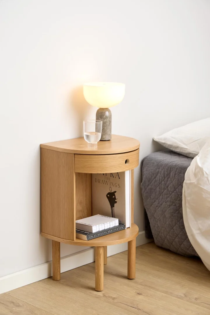Het Nachtkastje Eikenhout Slaapkamer, een modern houten nachtkastje met een open plank voor boeken en tijdschriften, staat bij een bed met daarop een grijs gewatteerde deken. Op het tafeltje staan een glas water en een lamp met een ronde, matte kap.