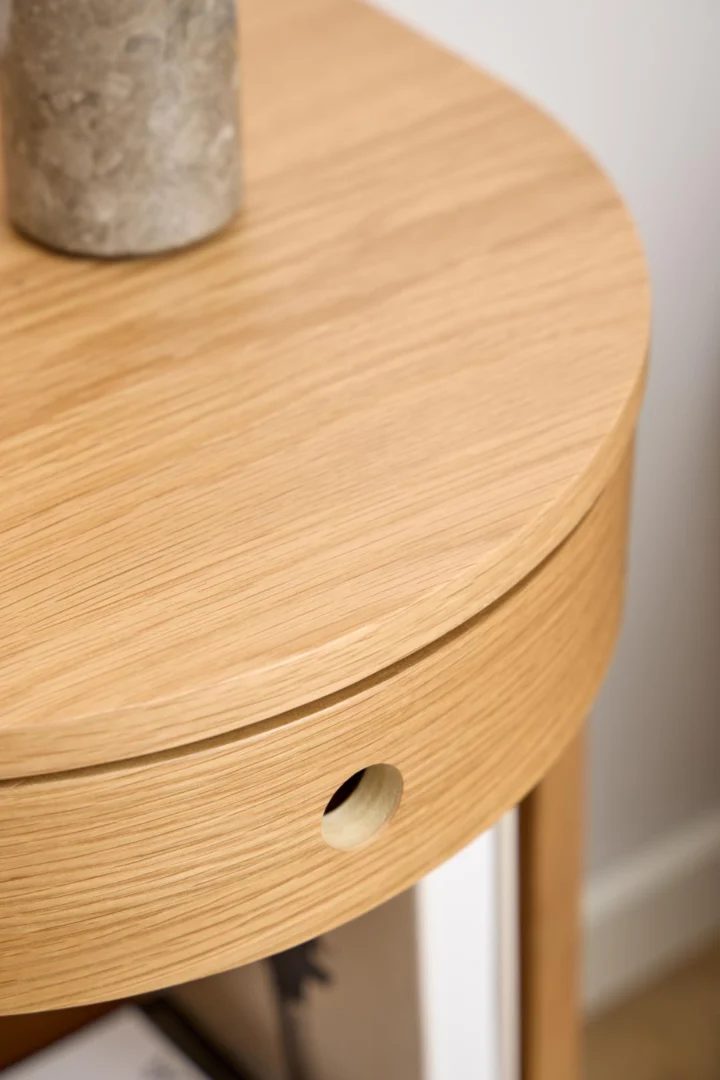 Close-up van het Nachtkastje Eikenhout Slaapkamer, een rond licht eiken nachtkastje met een zichtbaar handvatgat en een deel van een grijs stenen object bovenop, waardoor het natuurlijke houtnerfpatroon wordt benadrukt.