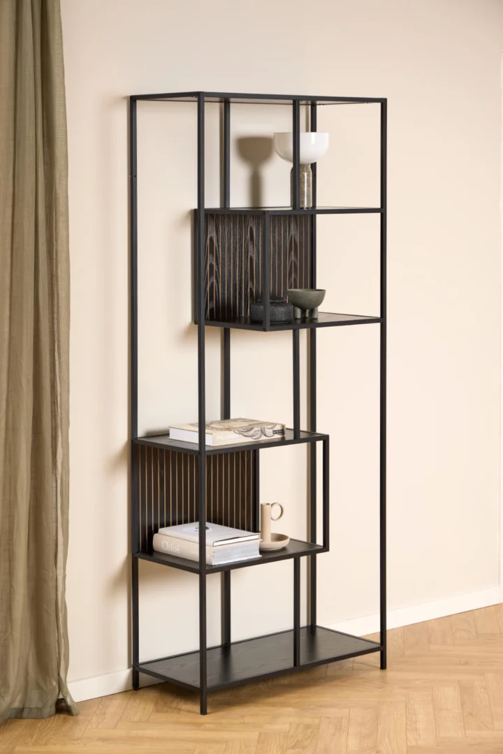 De Seaford boekenkast, een moderne open kast van zwart metaal en hout, staat tegen een beige muur met minimale decoratie zoals vazen, schalen, boeken en een mok; links verschijnt een groen gordijn.