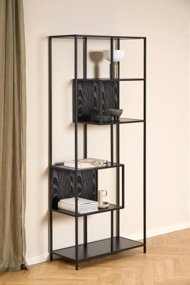 De Seaford boekenkast, een modern zwart metalen rek met open schappen, staat tegen een beige muur met boeken, een kom, twee kopjes en een mok. Links hangt een groen gordijn en op de vloer ligt licht houten visgraatmotief.