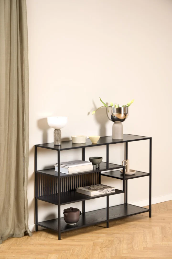 De Seaford boekenkast, een moderne zwarte metalen plank, toont vazen, schalen, boeken en keramiek tegen een crèmekleurige muur met houten vloer en een groenachtig gordijn aan de linkerkant voor een minimalistische inrichting.