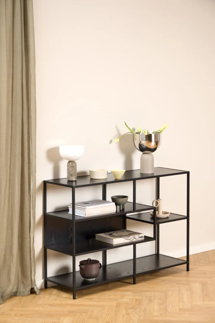 De Seaford boekenkast, een modern zwart metalen rek met boeken en vazen, staat tegen een beige muur met een lichte houten vloer en een groenachtig gordijn aan de linkerkant.