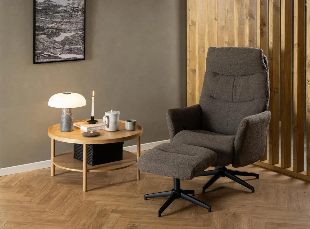 Een moderne woonkamer heeft een bruine fauteuil met bijpassende voetsteun, accenten van houten panelen, bruine muren, licht parket en een Salontafel Roest 80 CM Rond Eikenfineer die wordt gebruikt als bijzettafel met een lamp, kaars en theeservies.