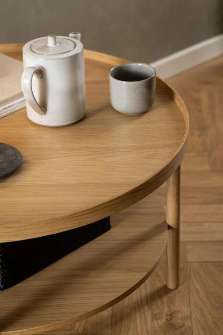 De Salontafel Roest 80 CM Rond Eikenfineer biedt plaats aan een theepot van keramiek, een bijpassende beker en een gesloten boek op het licht eikenhouten ronde blad. De salontafel heeft een onderste plank en staat op een parketvloer.