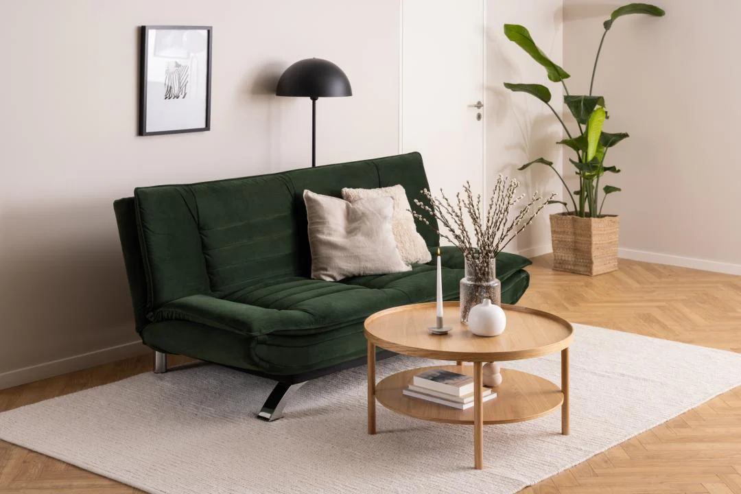 Een moderne woonkamer met een donkergroene bank, lichte kussens, een Salontafel Roest 80 CM Rond Eikenfineer met kaarsen en een vaas, een staande lamp, een potplant, muurkunst en een licht vloerkleed op een visgraat houten vloer.