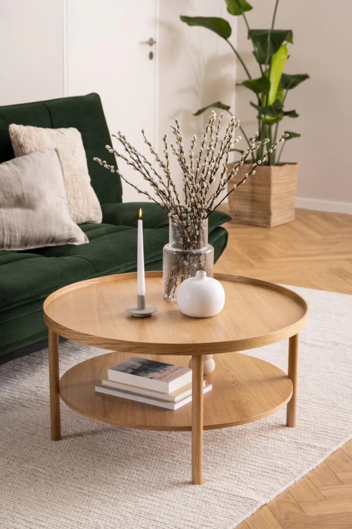 De Salontafel Roest 80 CM Rond Eikenfineer is ingericht met boeken, een witte vaas, een glazen pot met wilgentakken en een brandende kaars. Daarachter staan een groene bank met kussens, een grote potplant en een licht vloerkleed.