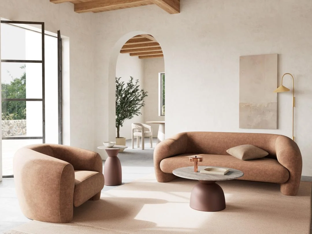 Een moderne woonkamer met een gebogen bruine bank en stoel, neutrale muurkunst, een gouden wandlamp en grote ramen. Het pronkstuk is de Salontafel Grijs, Brons 40 CM Rond in kunststof. Een boog leidt naar een eethoek met een plant.