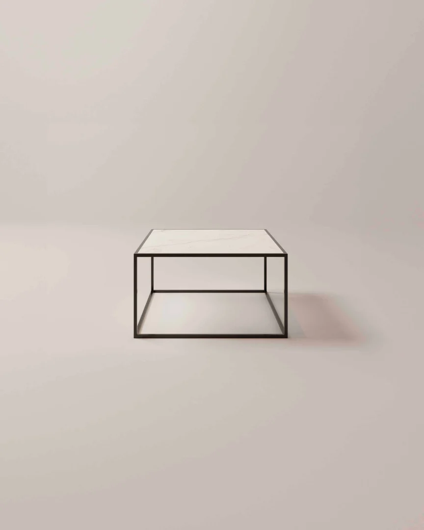 De Salontafel Zwart Klassiek Design is een minimalistische vierkante salontafel met een dun zwart metalen frame en een wit tafelblad tegen een effen lichtbeige achtergrond.