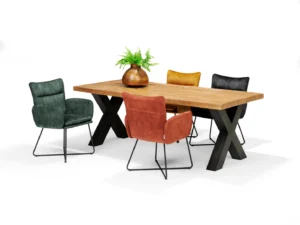 De Eettafel XILO Mango - 220x100, een houten eettafel met zwarte X-vormige poten, wordt gecombineerd met vier gecapitonneerde stoelen in groen, rood, geel en zwart. Een bruine vaas met groene varens decoreert de tafel tegen een witte achtergrond.