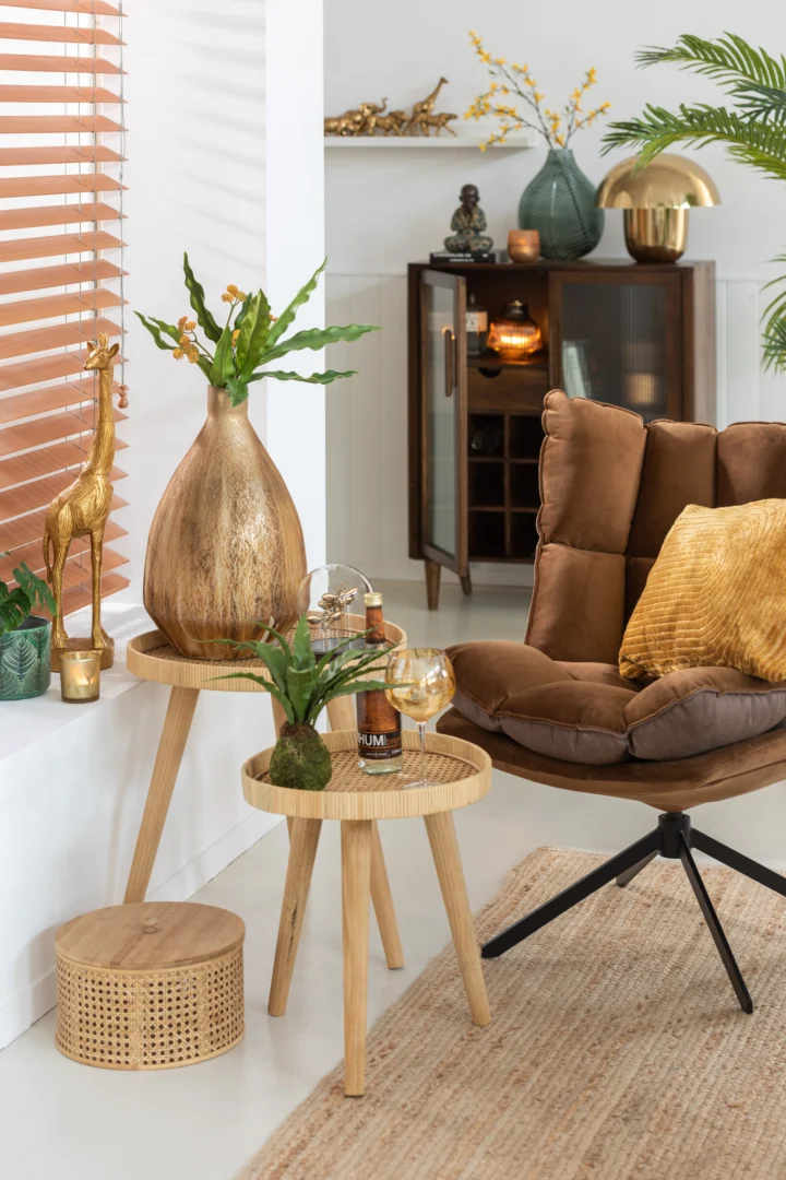 Een gezellige woonkamerhoek is voorzien van een bruine kussensstoel, gouden kussen, planten, een giraffesculptuur en twee Bijzettafels Rond Multiplex met vazen en drankjes. Warme natuurlijke tinten en een kast maken de look compleet.