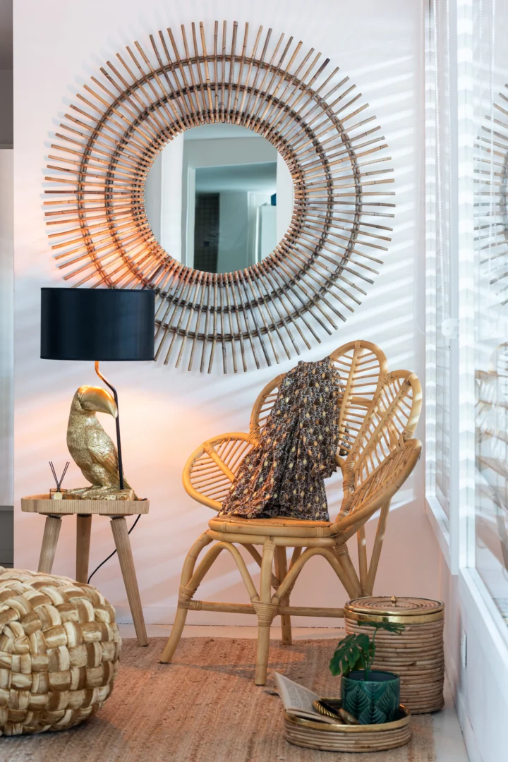Een knus hoekje met een ronde sunburst spiegel, een rotan stoel gedrapeerd met een gedessineerd kleed, Bijzettafel Rond Multiplex met een gouden vogellamp, geweven poef, groene plant, alles badend in natuurlijk licht.
