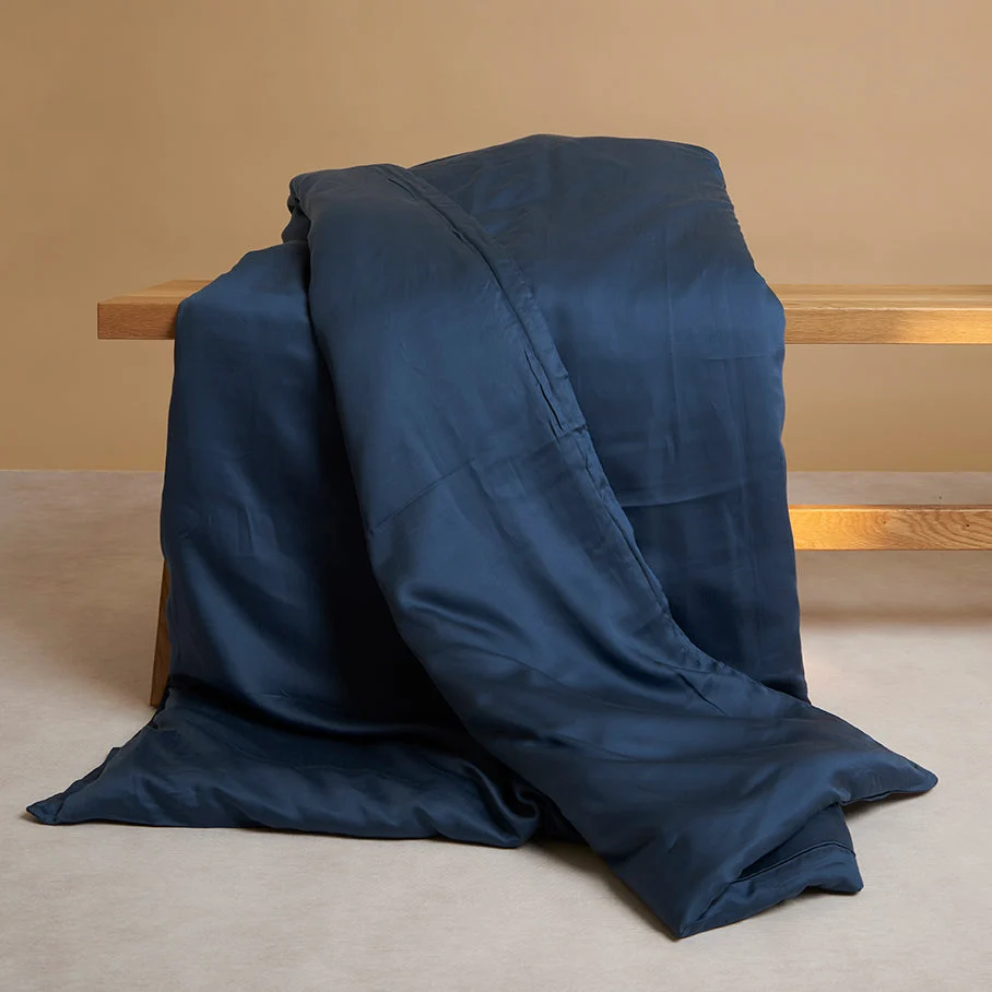 Blauw dekbedovertrek op houten bankje. De stof heeft een subtiele glans en valt in zachte plooien. Neutrale achtergrondkleuren.