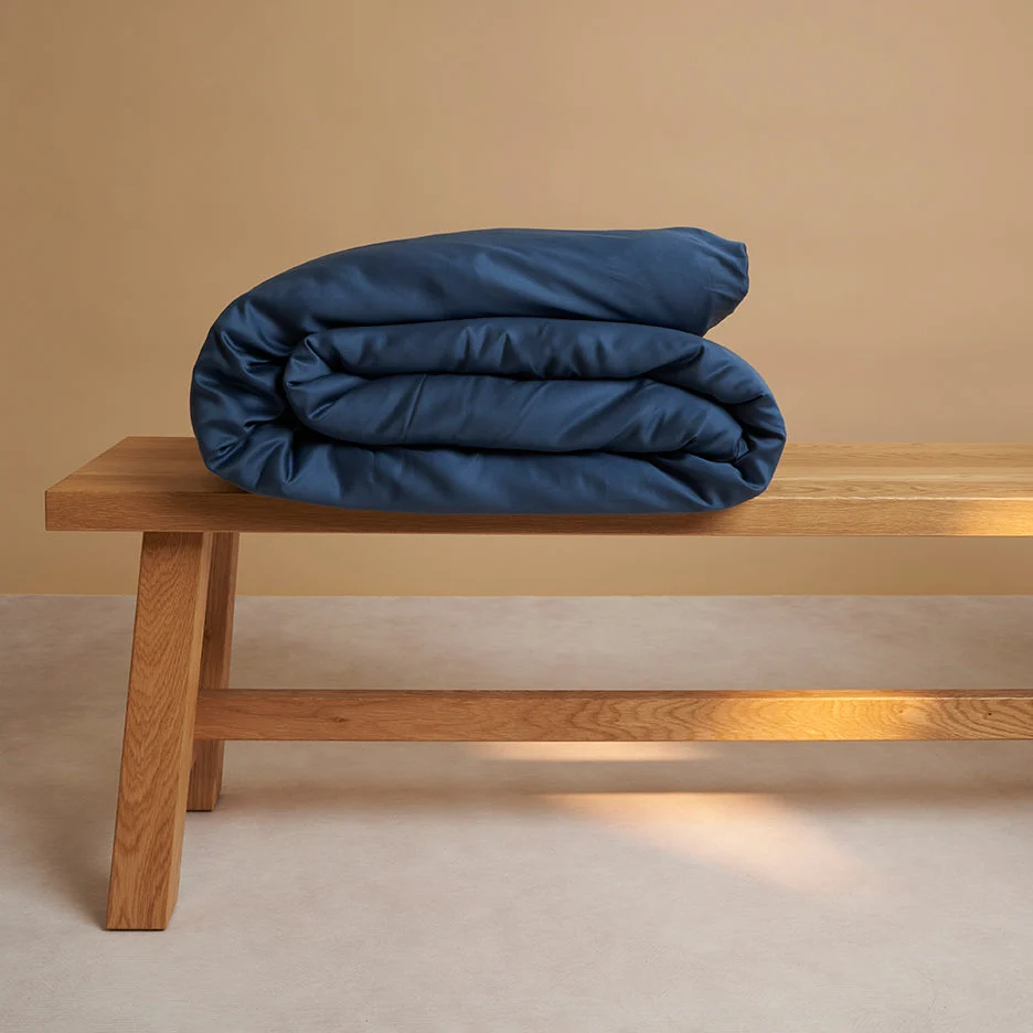 Gevouwen blauw dekbedovertrek op een houten bankje. Zachte belichting accentueert de textuur van het dekbed en het hout. Neutrale achtergrond.